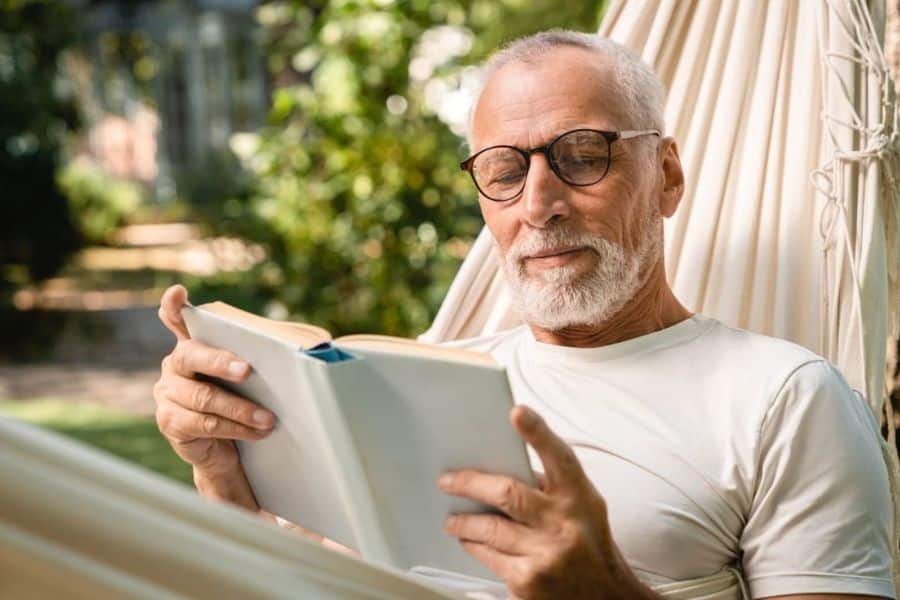 Older man with glasses and a white beard relaxes in an outdoor hammock while reading a book. Sunlight filters through the garden behind him giving the scene a quiet restful mood.