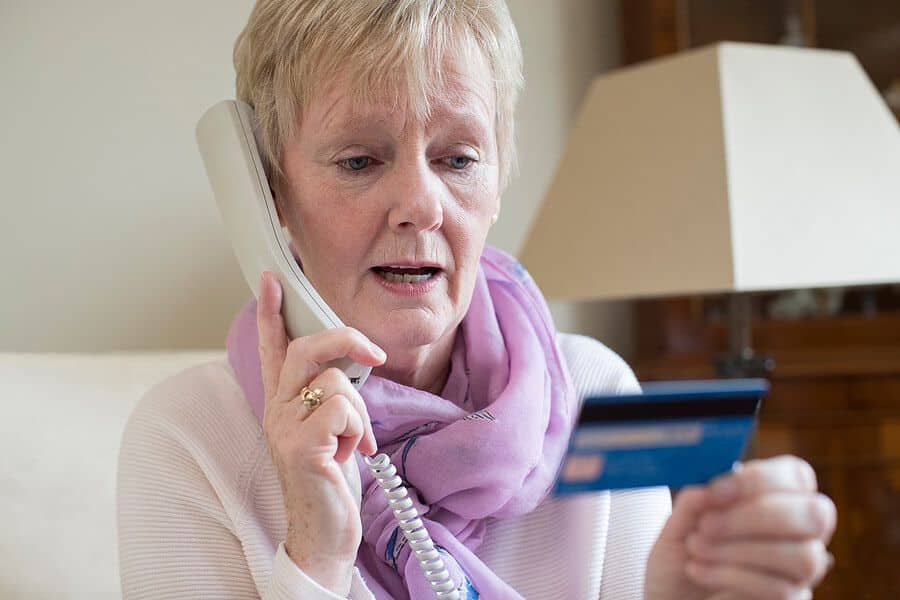 Older woman speaking on a landline while looking worriedly at a credit card in her hand. This scene shows how financial scams often target people through urgent phone calls involving payment or account details.