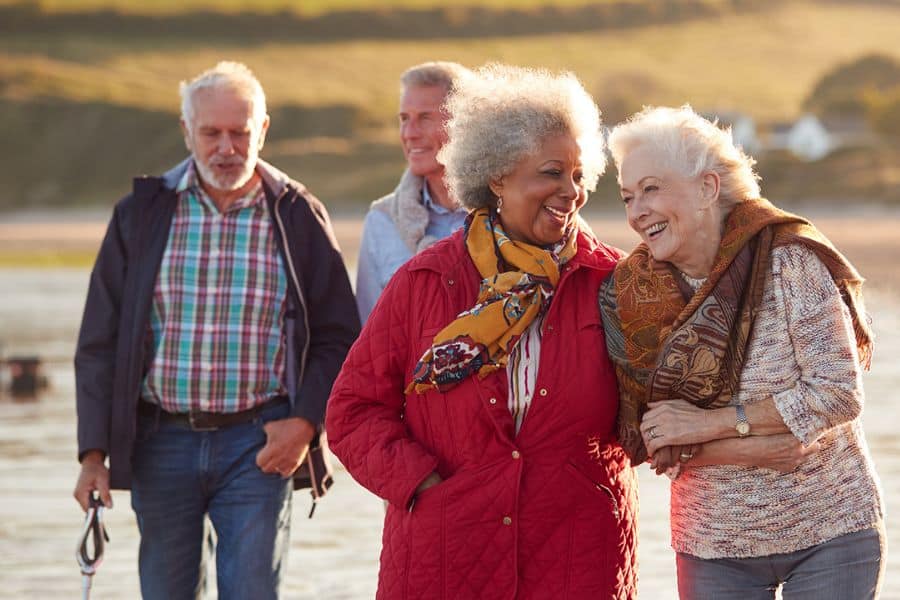 Group of older adults walking together along a beach or waterfront at golden hour, with two women in front smiling and linking arms. The photo represents friendship, activity, and companionship in later life.