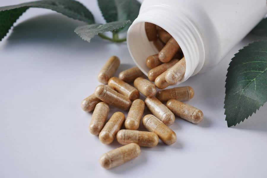 Open white supplement bottle tipped over with tan capsules spilled onto a white surface beside green leaves. The photo represents supplements that may be discussed in relation to chronic inflammation.