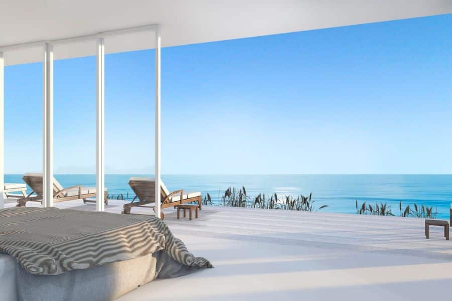 Minimal bedroom with floor to ceiling glass doors opening onto a deck with lounge chairs and an uninterrupted view of the blue sea and sky. The peaceful setup evokes an affordable dream getaway for people hoping to see the world on a budget.