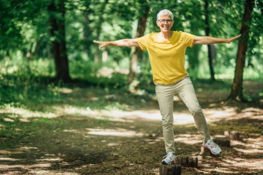 Older woman in a yellow shirt balances with her arms stretched out while standing on wooden posts along a forest path. The image highlights balance training and confidence outdoors.
