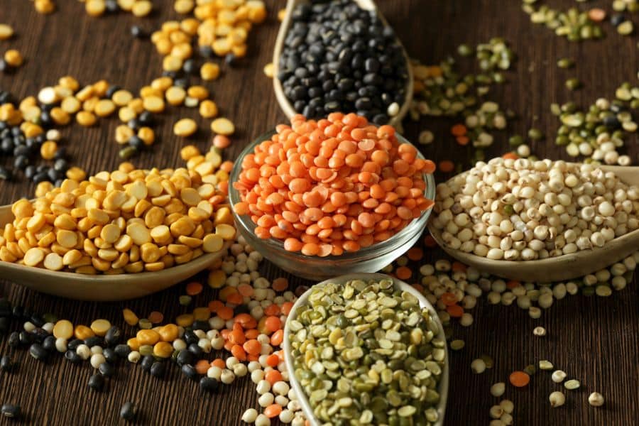 Assorted dried legumes and pulses scattered across a dark wooden surface, with red lentils in a glass bowl and black beans, split peas, yellow peas, and white sorghum arranged in wooden spoons around them. This ingredient spread supports content about artery plaque by showing fiber rich plant foods often associated with heart healthy eating.