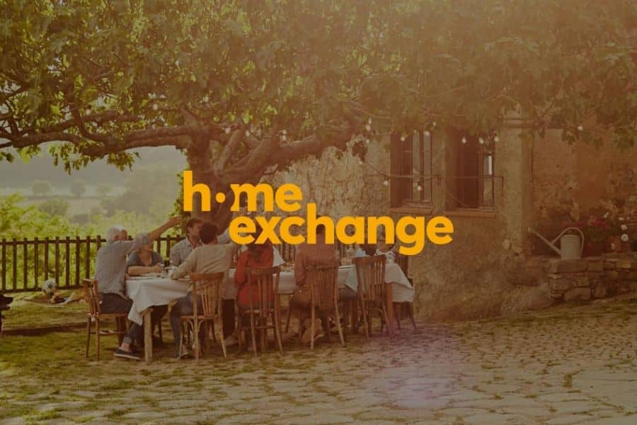 Outdoor dinner gathering under a large tree beside a rustic stone house, with the words "h.me exchange" centered in bold yellow text over the scene. The image points to home exchange as a social and cost saving way to see the world on a budget.
