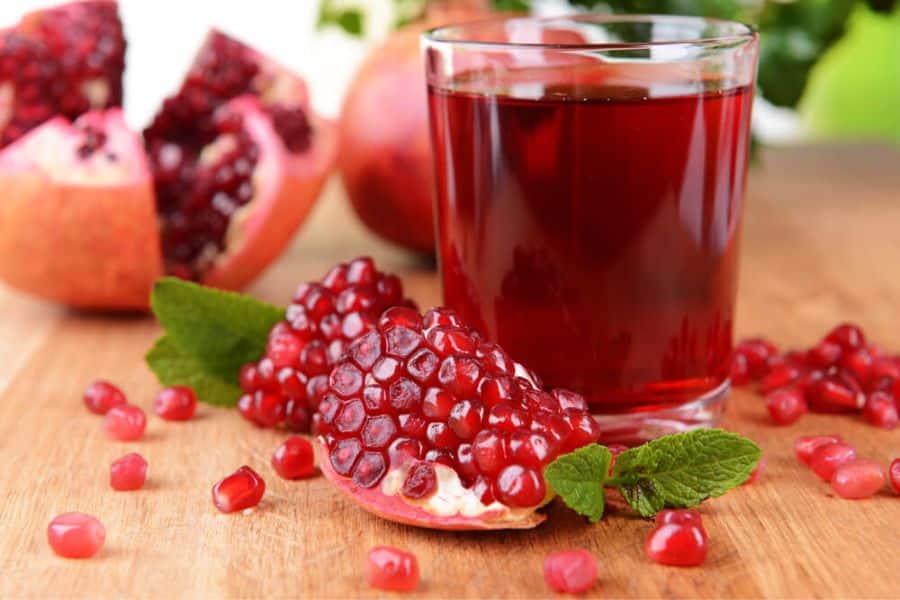 Glass of deep red pomegranate juice on a wooden table with opened pomegranates, loose seeds, and fresh mint leaves around it. This photo fits content about artery plaque by highlighting pomegranate as a fruit commonly featured in heart health discussions.