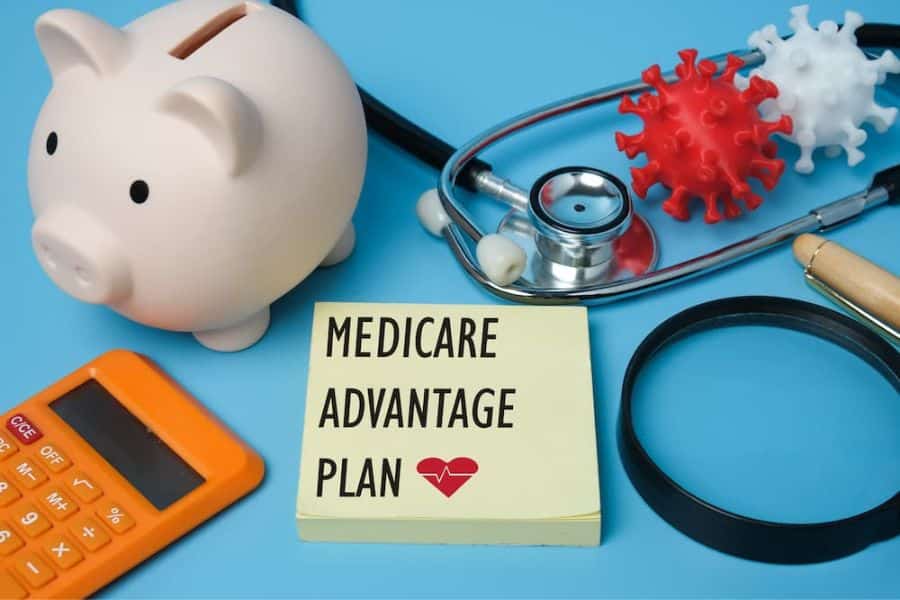 A piggy bank, calculator, stethoscope, magnifying glass, and virus models surround a note that reads "MEDICARE ADVANTAGE PLAN." The composition highlights Medicare benefits related to healthcare costs, plan choices, and medical coverage.