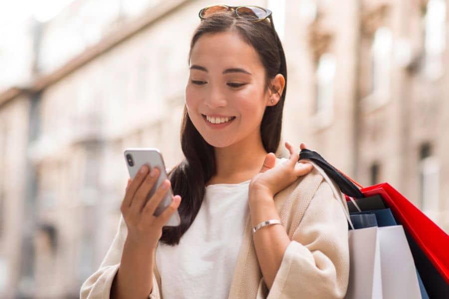 Woman outdoors smiles at her phone while carrying several shopping bags over one shoulder. This works for retailer tricks content about mobile promotions, app alerts, and limited time offers that encourage extra spending.