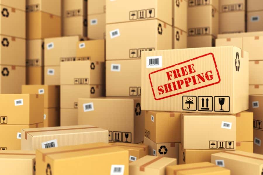 Stacks of cardboard shipping boxes fill a warehouse scene, with one large box stamped with the words "FREE SHIPPING". This supports retailer tricks content by showing how free shipping offers are used to make online deals feel more valuable.