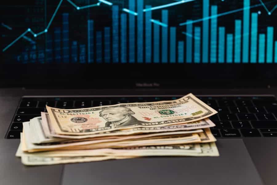 A stack of US dollar bills sits on a laptop keyboard in front of a blue market chart on the screen. This visual supports content about returns performance and building wealth through index funds.
