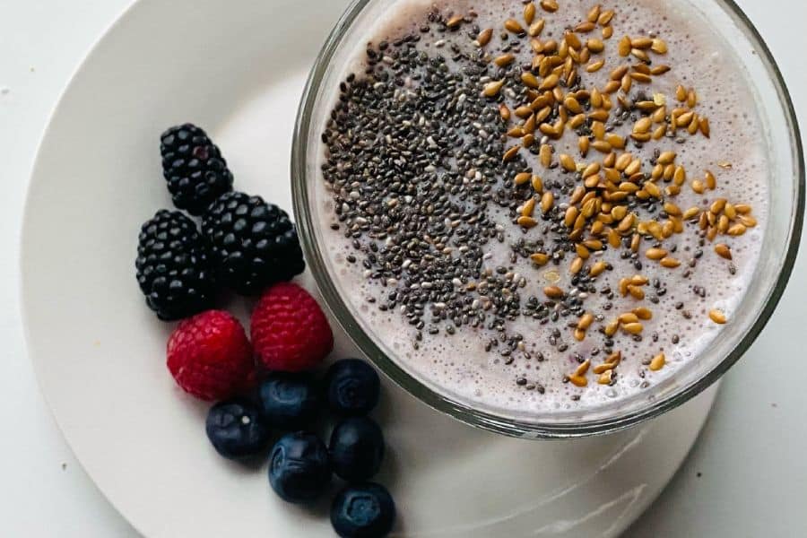 Creamy smoothie in a glass bowl topped with chia seeds and flaxseeds, served beside blackberries, raspberries, and blueberries on a white plate. This breakfast style dish relates naturally to artery plaque content by showing berries and seeds often included in heart conscious meals.