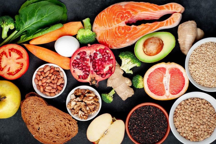 A flat lay of salmon, avocado, pomegranate, grapefruit, lentils, grains, nuts, bread, and fresh vegetables arranged on a dark surface. The colorful spread connects to Scandinavian lifestyle habits that often emphasize wholesome ingredients and balanced meals.