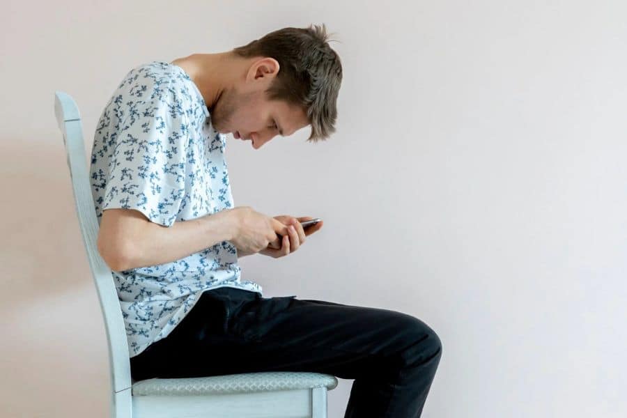 Young man sits on a chair while looking down at a phone with his upper back rounded and neck bent sharply forward. This posture shows the slouched position commonly associated with prolonged phone use.