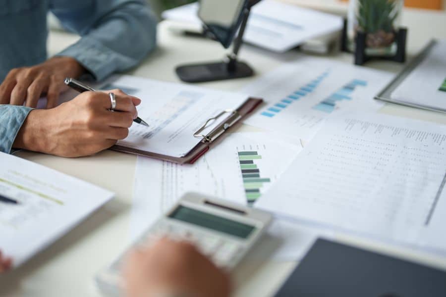 Close up of a person reviewing financial documents with charts and graphs while writing notes on a clipboard at a desk with a calculator nearby. The scene shows the hands on process behind decisions when people use AI to plan your retirement alongside traditional financial analysis.