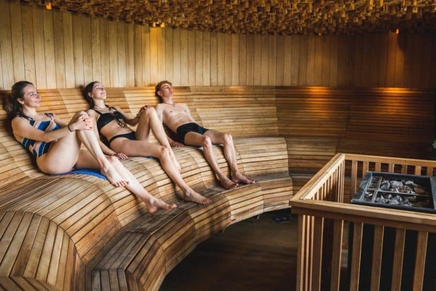 Three people relax on curved wooden benches inside a softly lit sauna with warm paneled walls. The sauna scene highlights Scandinavian lifestyle habits that prioritize wellness, rest, and daily rituals of recovery.