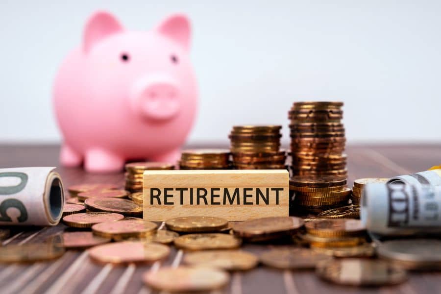 Wooden block labeled "RETIREMENT" sits among scattered coins and rolled banknotes, with a pink piggy bank and stacked coins blurred in the background. This photo clearly illustrates retirement saving and building a retirement fund over time.