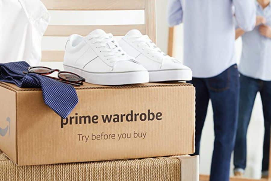 Cardboard box topped with white sneakers, glasses, and folded clothing sits in the foreground while two people stand blurred in the background. The box text reads "prime wardrobe" and "Try before you buy", which makes it a strong fit for retailer tricks content about low risk trial offers that encourage more ordering.
