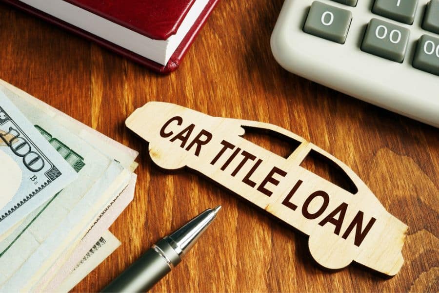 Wooden car shaped sign reading "CAR TITLE LOAN" sits on a desk beside cash a pen a book and a calculator. This image illustrates borrowing money using a vehicle title as collateral.