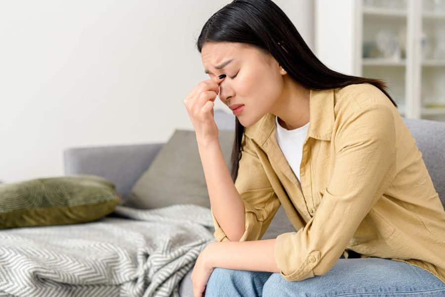 Woman sitting on the edge of a bed with her eyes closed and fingers pressed to the bridge of her nose in visible stress or frustration. This reflects destructive habits tied to emotional exhaustion and feeling overwhelmed.