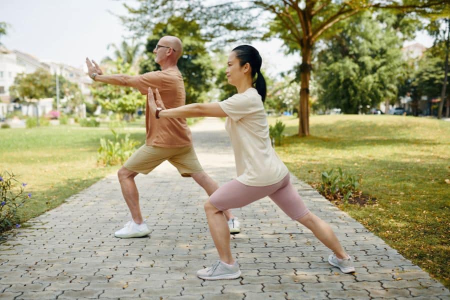 Two adults exercise on a paved park path with arms extended and knees bent in a balanced pose. The outdoor workout scene supports movement and gentle exercise for chronic inflammation.