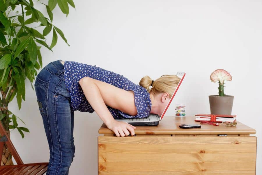 Blonde woman bent over a wooden desk with her face pressed into an open laptop in a humorous but exaggerated pose of burnout. The cluttered workspace reinforces destructive habits linked to overwork and mental fatigue.