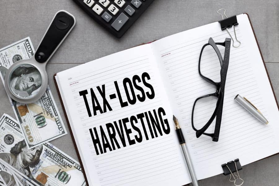 Notebook with bold text "TAX-LOSS HARVESTING" surrounded by cash glasses and a calculator illustrating an investment strategy for paying less tax. Close up view emphasizes financial tools and planning details.