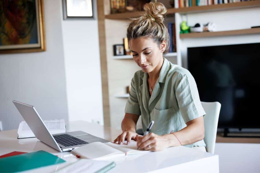 A woman sits at a desk writing in a notebook beside an open laptop in a home office. The scene suggests researching and tracking a simple investment plan such as index funds.