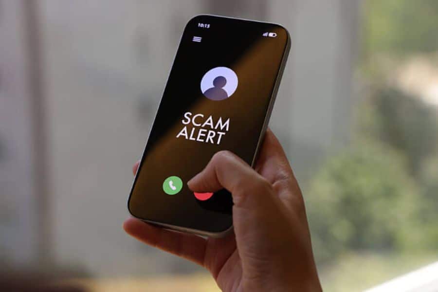 Hand holding a smartphone showing an incoming call screen with the words "SCAM ALERT." This image emphasizes financial scams delivered through fraudulent phone calls.