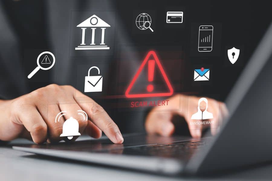 Person using a laptop with digital security icons floating above the keyboard including a red warning symbol labeled "SCAM ALERT" and a small figure labeled "RANSOMEWARE." This image highlights online financial scams and cyber threats linked to banking and personal data.
