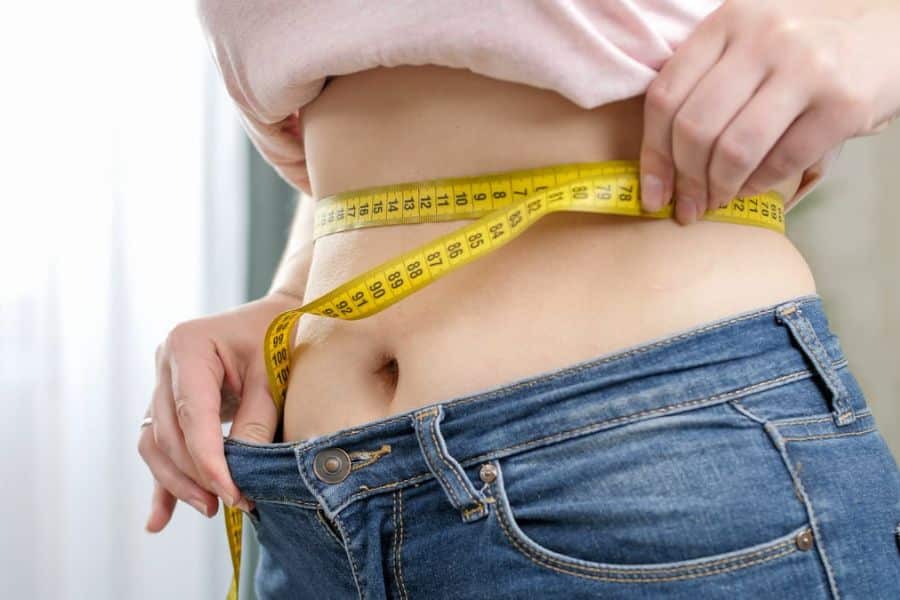 Close up of a person measuring their waist with a yellow tape measure while pulling out the waistband of loose blue jeans. The photo visually represents weight loss or changes in body size.