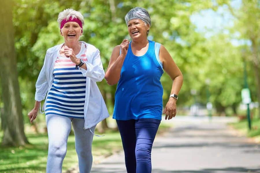 Two older women in athletic clothes walk side by side on a tree lined path, smiling and talking as they exercise together. The scene shows walking for fitness, health, and social support in older adulthood.