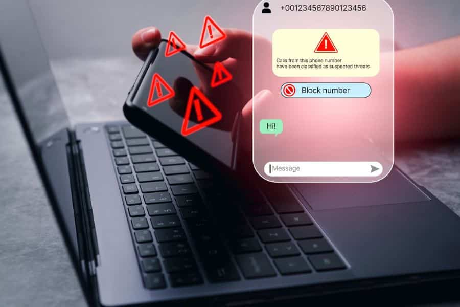 Hand holding a phone over a laptop with red warning icons and a message screen showing a suspected threat. The screen says "+001234567890123456" "Calls from this phone number have been classified as suspected threats." "Block number" "Hi." and "Message."