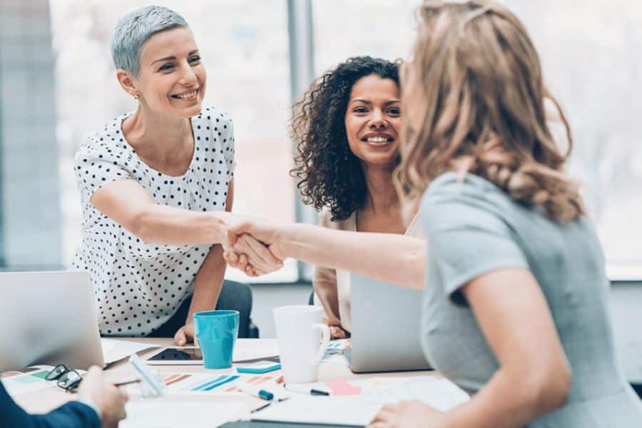 Three women sit around a meeting table while two of them shake hands and smile. This photo fits Medicare benefits content about getting help from an advisor or support team when choosing coverage.