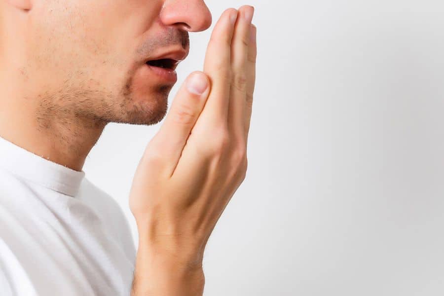 Side view of a man in a white shirt raising his hand to his mouth as if checking unpleasant breath. The plain background keeps the focus on possible bad breath as a symptom.