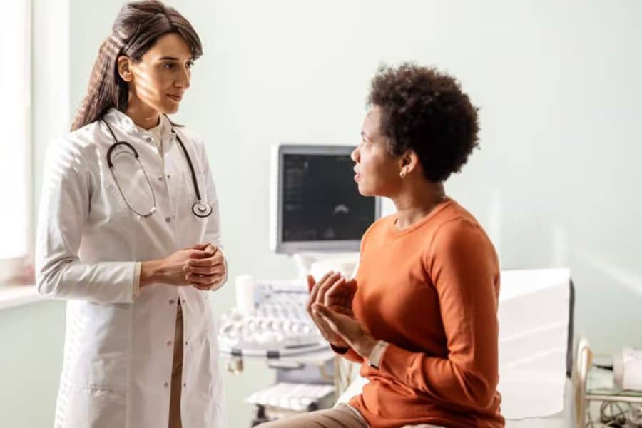 A doctor with a stethoscope speaks with a woman seated on an exam table in a medical office. This supports Medicare benefits by showing a routine doctor visit and patient care conversation.