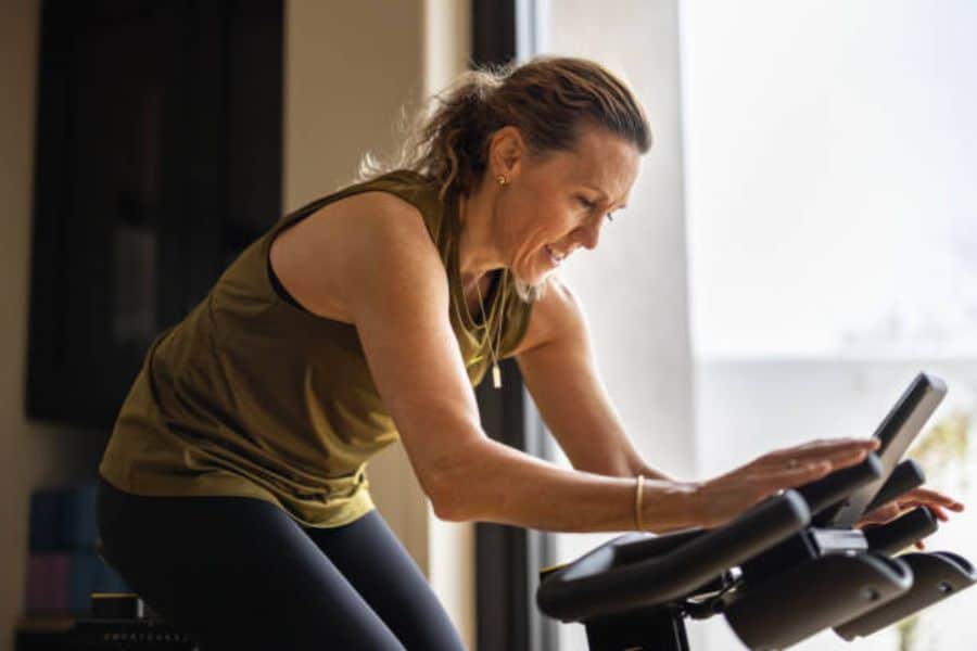 Woman exercising on a stationary bike indoors while adjusting the workout screen and smiling during her cardio session. The image highlights healthy habits by showing a consistent at home fitness routine that supports an active lifestyle.