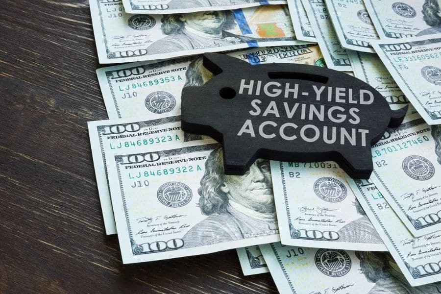 Black piggy bank shaped sign labeled “HIGH-YIELD SAVINGS ACCOUNT” placed on top of scattered one hundred dollar bills. The image represents building an emergency fund through high interest savings and long term financial security.