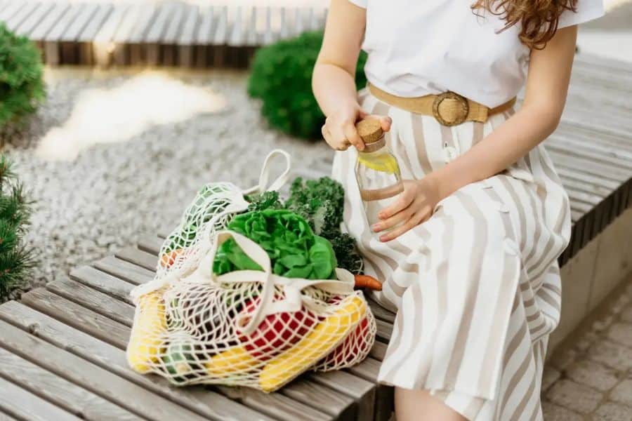 Woman sitting outdoors beside reusable mesh grocery bags filled with fresh vegetables while holding a reusable water bottle. The scene highlights sustainable switches like low waste shopping, reusable containers, and buying fresh produce locally.