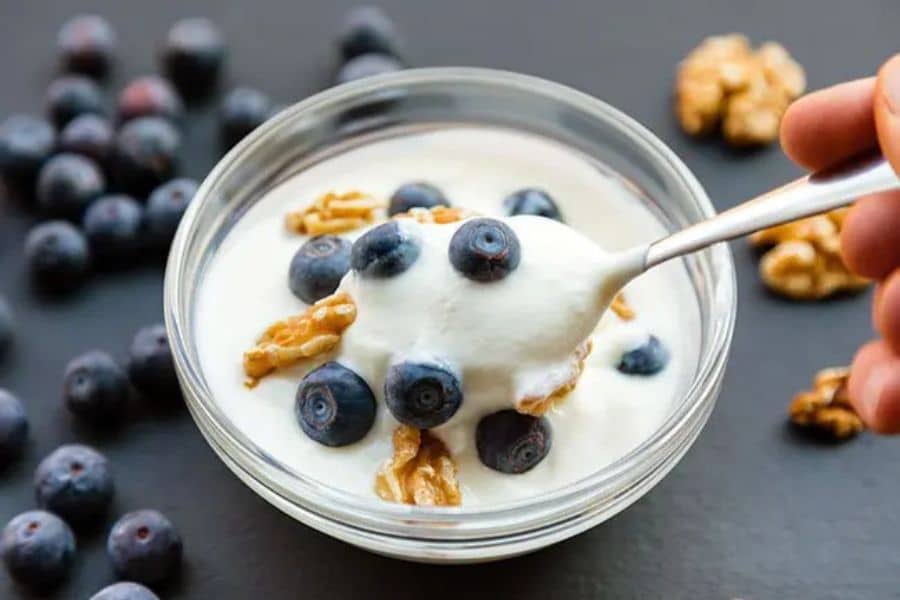 Bowl of yogurt topped with fresh blueberries and walnuts with a spoon lifting a serving from the bowl. This nutritious snack supports healthy habits through balanced ingredients rich in protein, fruit, and healthy fats.