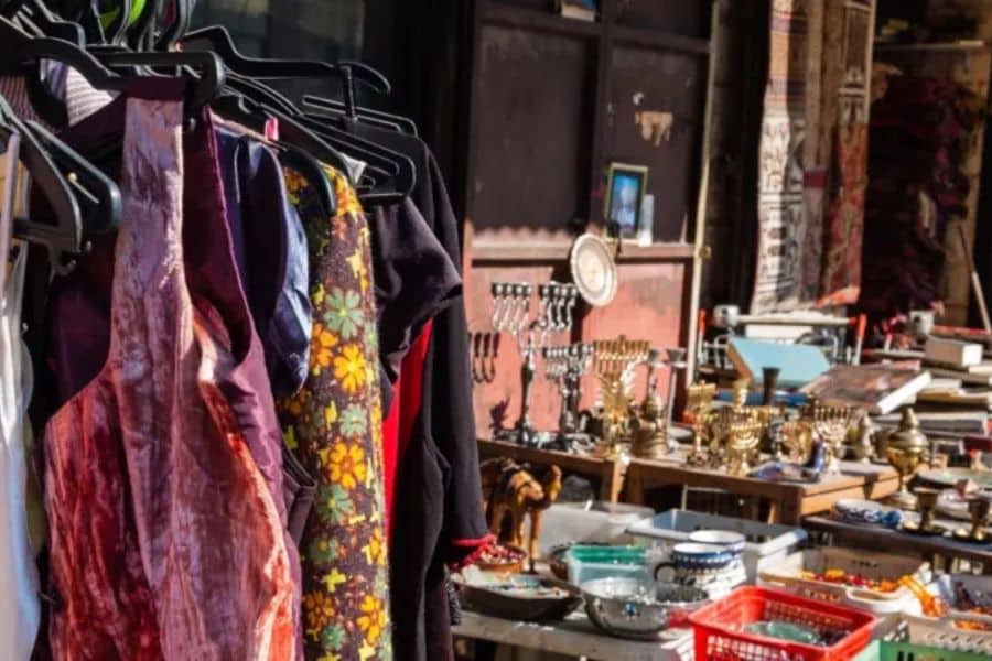 Clothing racks and secondhand household items displayed at an outdoor flea market with dresses, dishes, books, and vintage decor visible in the background. The scene relates to saving money for an emergency fund through budget friendly shopping and reducing unnecessary spending.