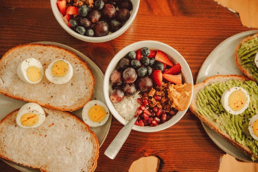 Breakfast table with avocado toast topped with boiled eggs, bowls of mixed berries and grapes, and yogurt with fruit and granola. The colorful spread represents healthy habits through balanced meals made with fresh and nutrient dense ingredients.