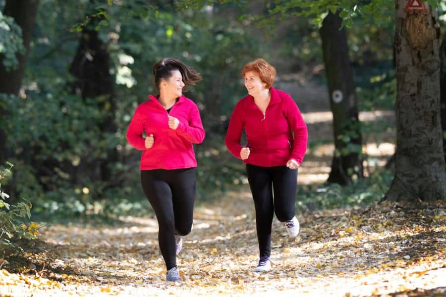 Two women jogging together along a wooded trail while wearing matching pink athletic jackets and black leggings. The outdoor exercise scene reflects healthy habits through regular movement and social fitness activities.