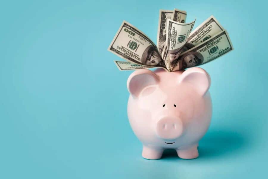 Pink piggy bank filled with folded dollar bills against a light blue background. The simple savings image symbolizes setting aside money for an emergency fund and future financial goals.