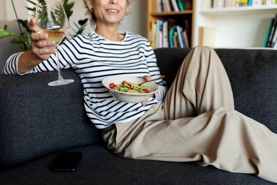 An older woman with a glass of white wine and a bowl of food sat on a couch