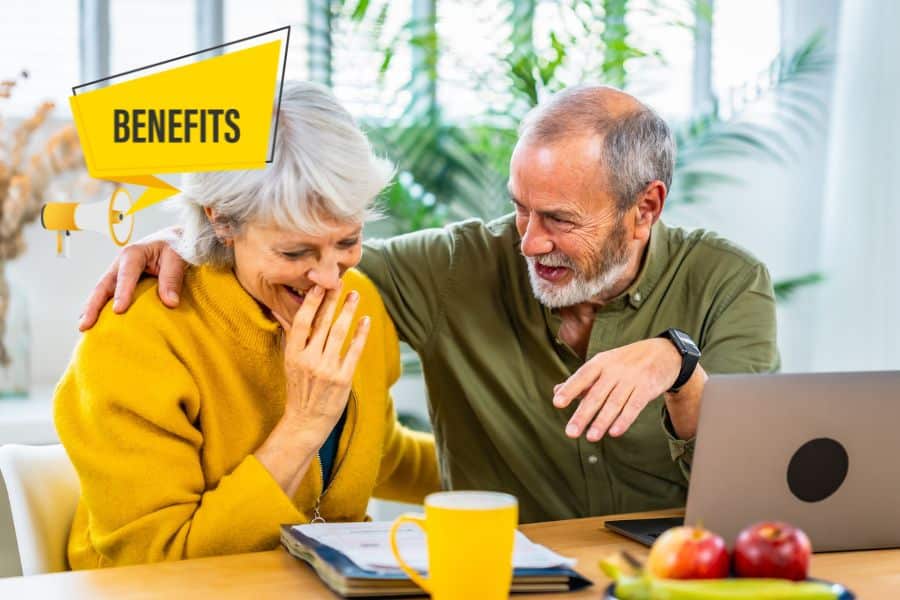 Older couple laughing together at a table with a laptop and a yellow sign that says "BENEFITS." They appear to be reviewing Medicare benefits in a bright home office.
