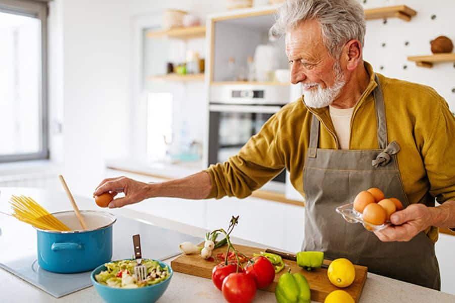 Older man preparing a homemade meal in a bright kitchen while holding eggs beside fresh vegetables, lemons, and a bowl of salad ingredients. The healthy cooking scene supports a superfoods lifestyle through fresh ingredients, balanced nutrition, and home prepared meals.