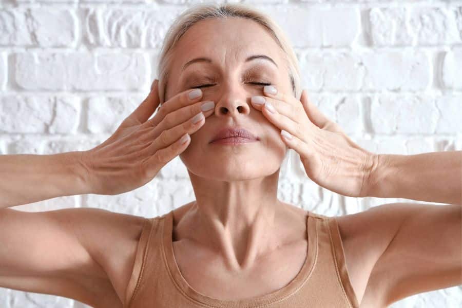 Woman with closed eyes gently massages the skin beneath her eyes with both hands while practicing face yoga against a white brick wall. The close up pose highlights relaxing facial movements often used in face yoga routines for circulation and muscle tone.