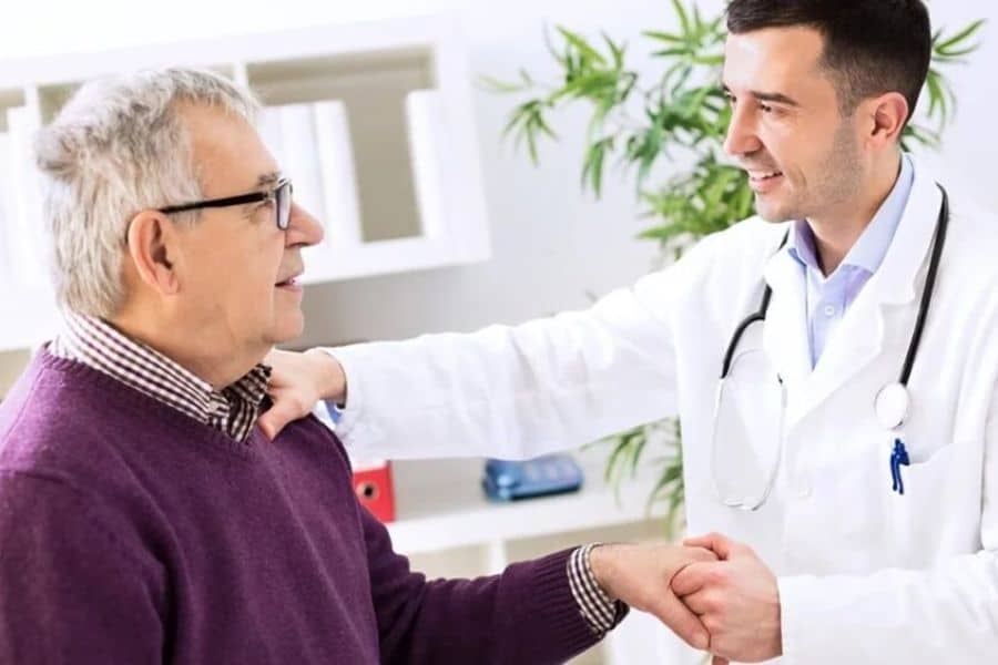 Doctor smiling and holding hands with an older patient during a visit. The friendly appointment setting suggests support with Medicare related care.