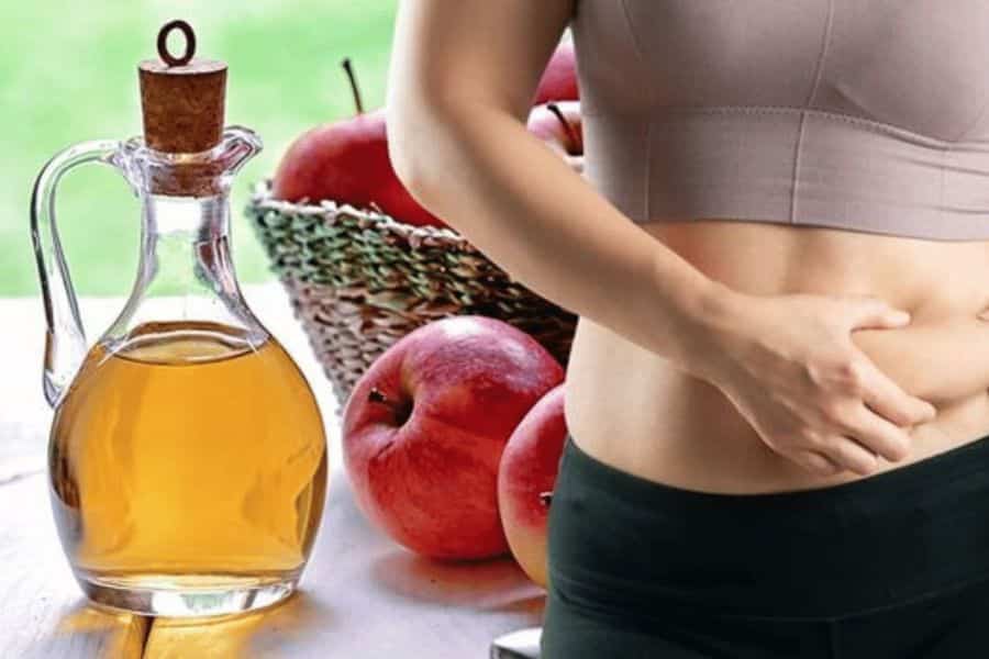Glass bottle of apple cider vinegar beside red apples and a woman holding her stomach while wearing workout clothes. The image suggests apple cider vinegar as part of a wellness or digestive health routine.