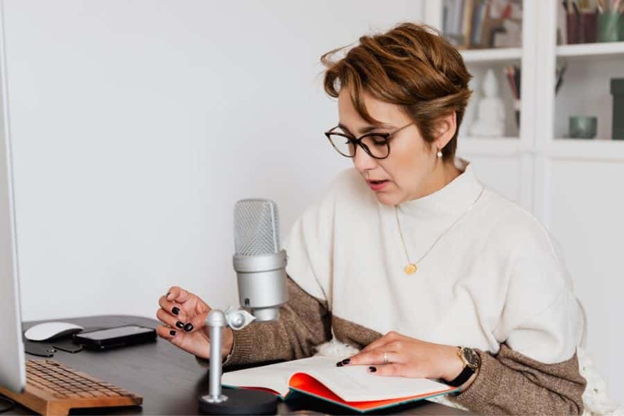 Woman wearing glasses speaking into a microphone while reading notes at a desk with a computer nearby. She appears to be recording a podcast or online content as part of building income streams.