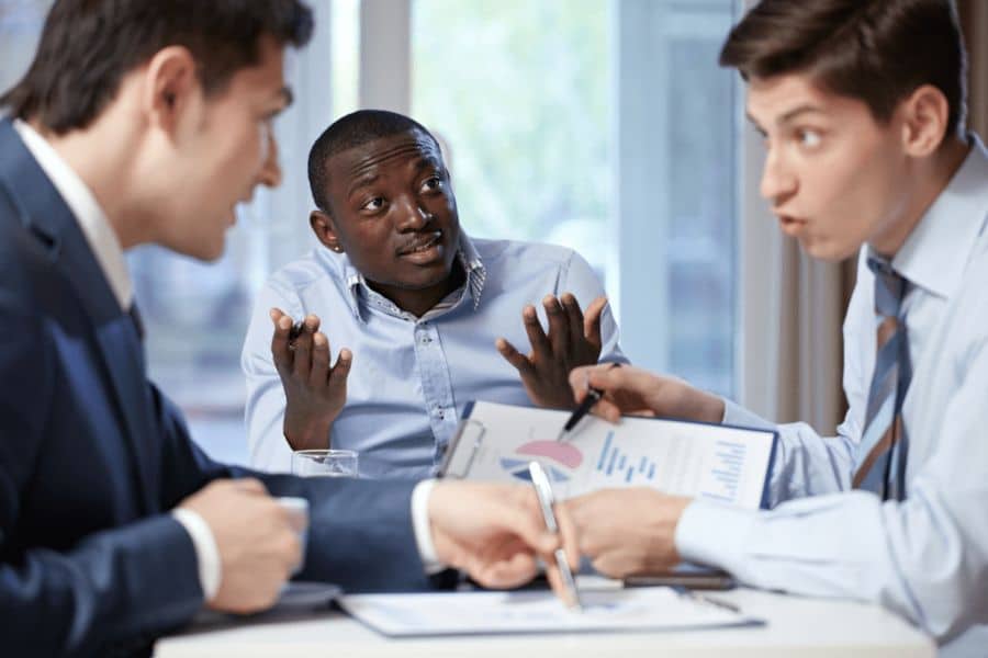 Three business professionals seated around a table during a discussion with charts and documents spread out in front of them. The meeting suggests teamwork and strategy planning for new income streams.